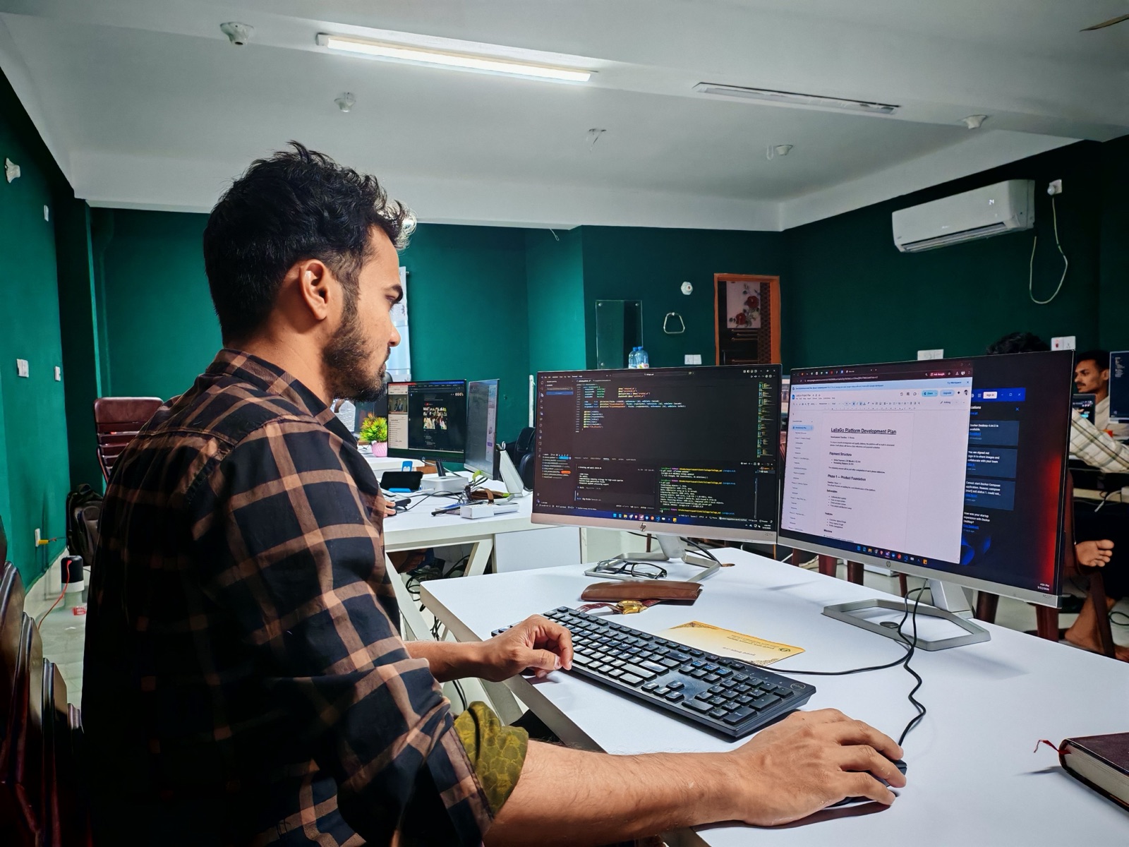 A man in a plaid shirt working at a desk with two monitors