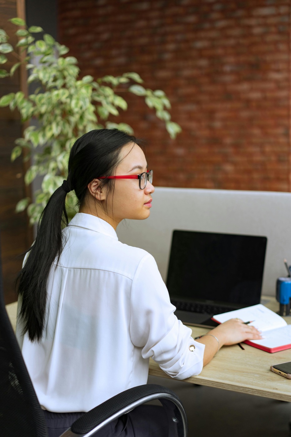 A person with red glasses taking notes at a desk beside a laptop